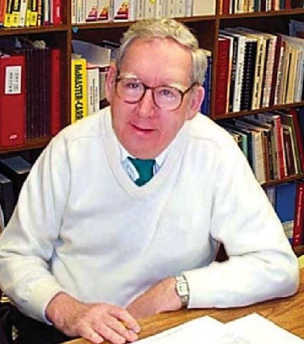 Albert E. Sanderson seated at a desk in front of bookshelves, wearing glasses and a white sweater
